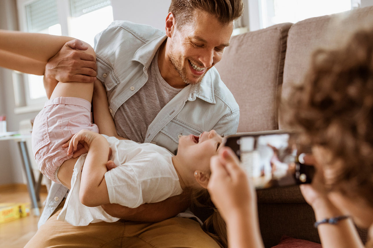 Vater und Kind albern im Wohnzimmer und werden dabei von dritter Person mit Handy fotografiert