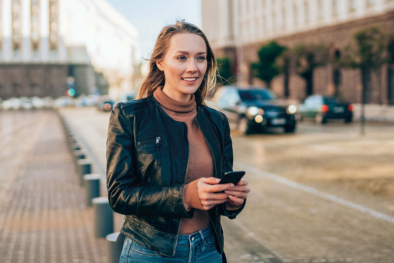 Junge Frau mit Smartphone in der Hand geht auf der Straße und lächelt