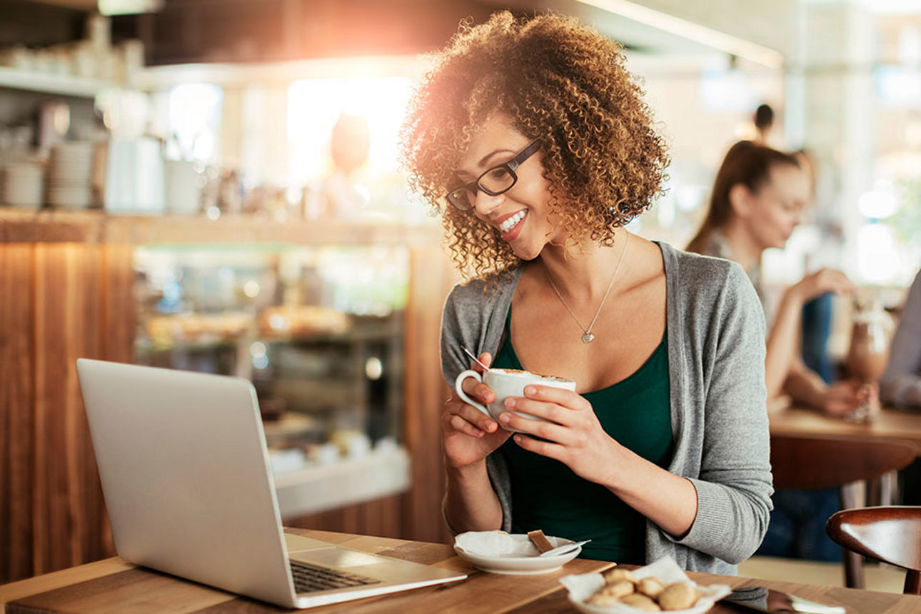 Frau mit lockigen Haaren sitzt im Café und schaut lächelnd auf Ihren Laptop