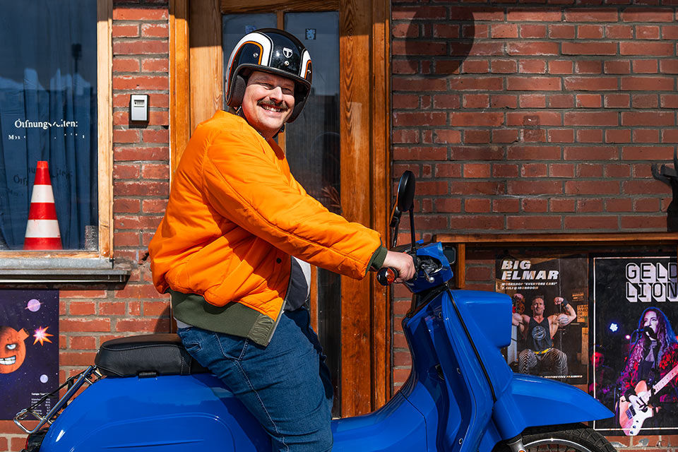 Mann mit Helm und orangenfarbener Jacke sitzt auf einem blauen Moped und lächelt in die Kamera