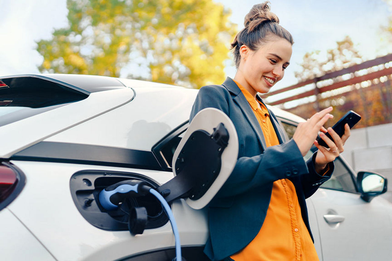 Eine junge Frau lehnt sich beim Tanken an ihr Auto und schaut auf ihr Handy