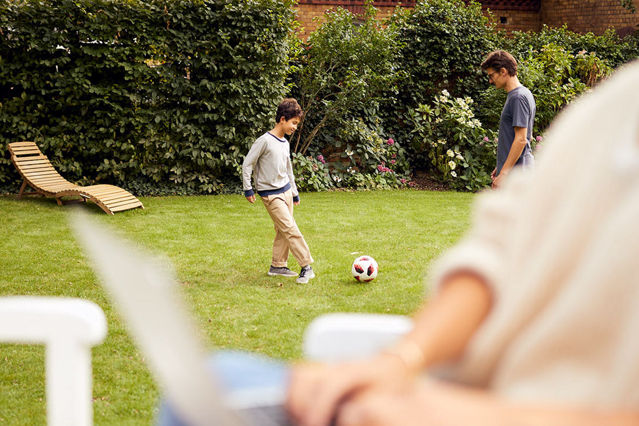 Eine Frau sitzt mit Laptop im Garten, während ihr Sohn und ihr Mann Fußball spielen