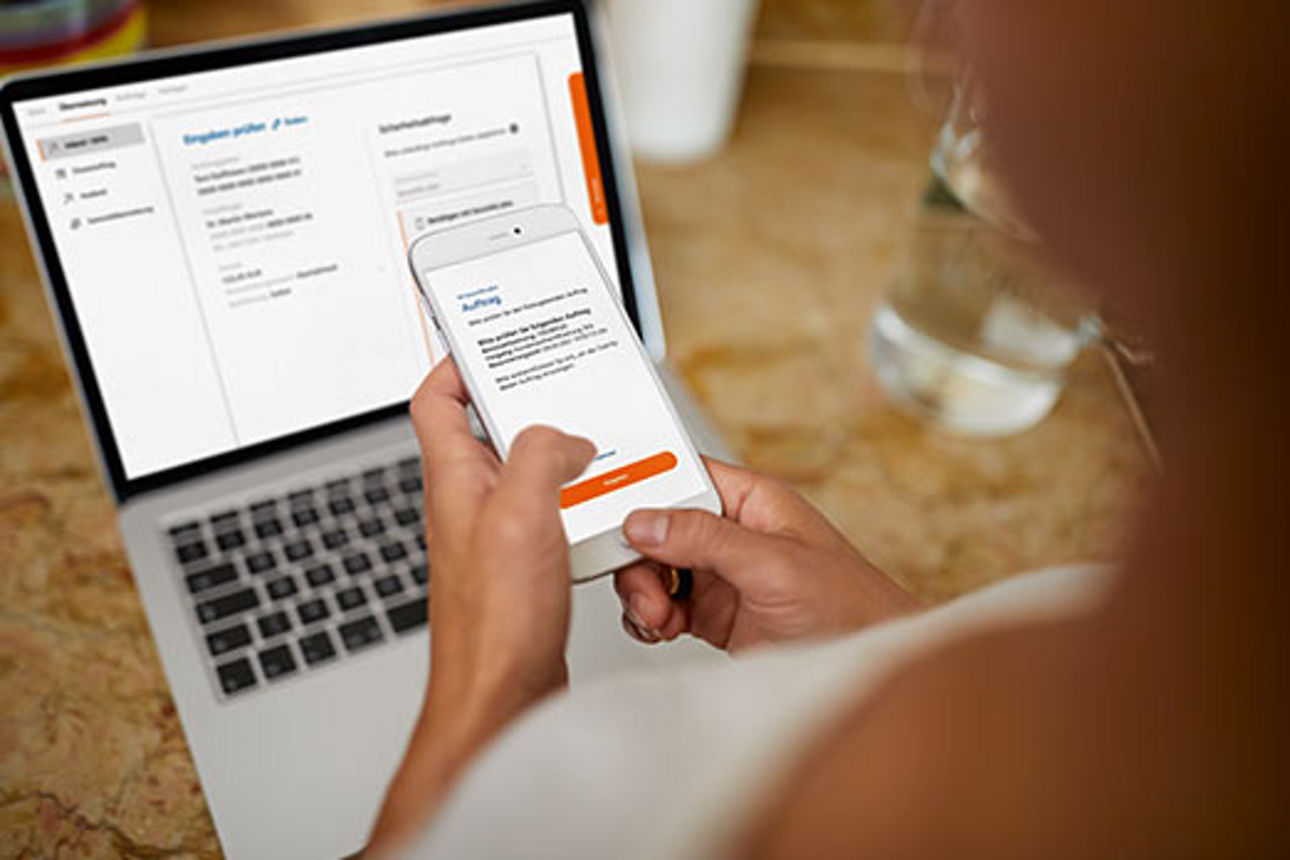 Person vor einem Laptop, mit einem Smartphone in der Hand, auf dem eine TAN-App geöffnet ist